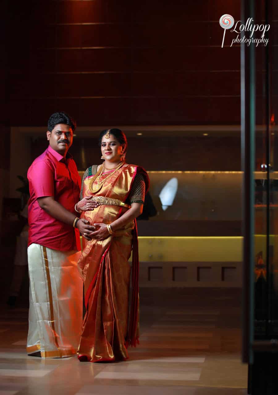 A couple stands together during a baby shower event in Chennai; the pregnant woman in a richly adorned saree holds her belly, standing next to her partner in vibrant attire