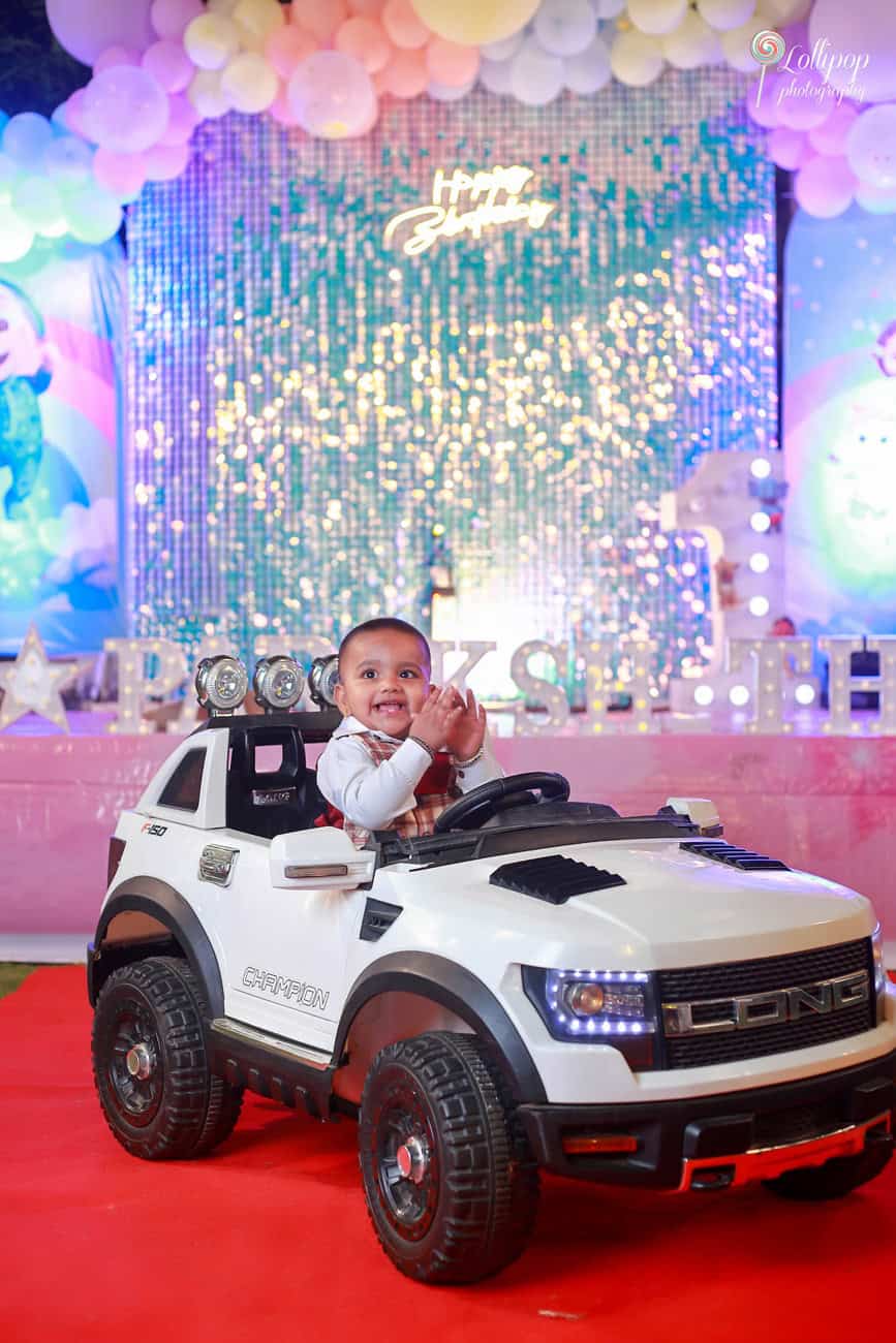 A little boy, smiling joyously, sitting in a white toy SUV, waving at a birthday party in Chennai, with a magical sequined backdrop enhancing the festive mood.