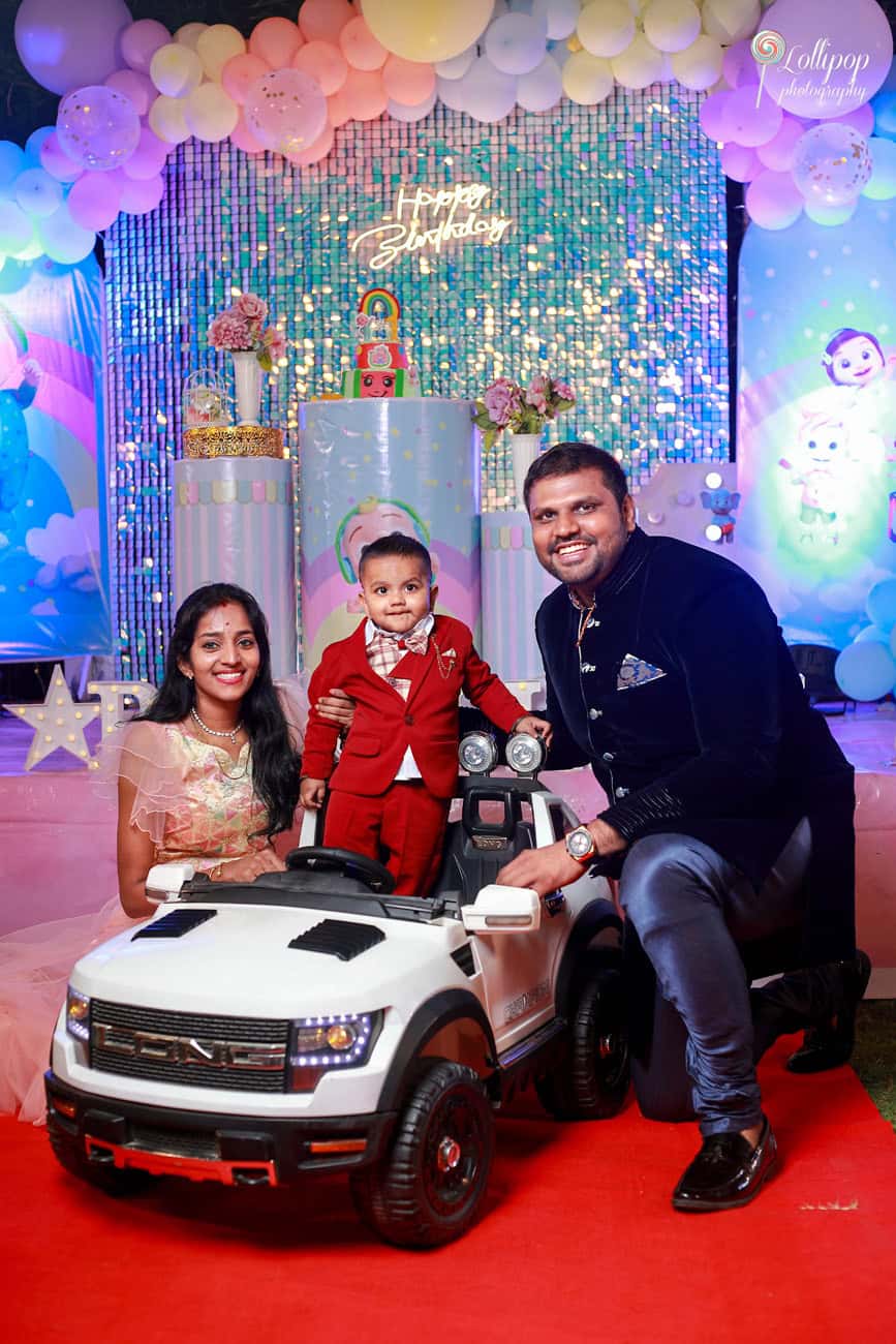 A charming family poses with their child in a red toy car at a birthday party, surrounded by colorful balloons and a glowing "Happy Birthday" backdrop in Chennai.