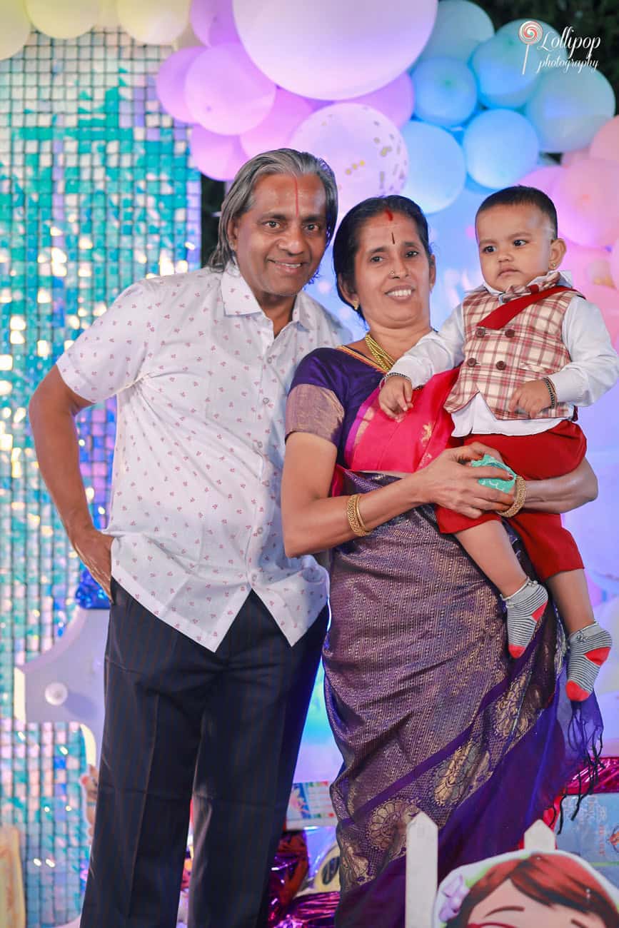 Grandparents celebrating their grandchild's birthday at a party in Chennai, with a cake cutting ceremony in front of a twinkling blue sequined backdrop.