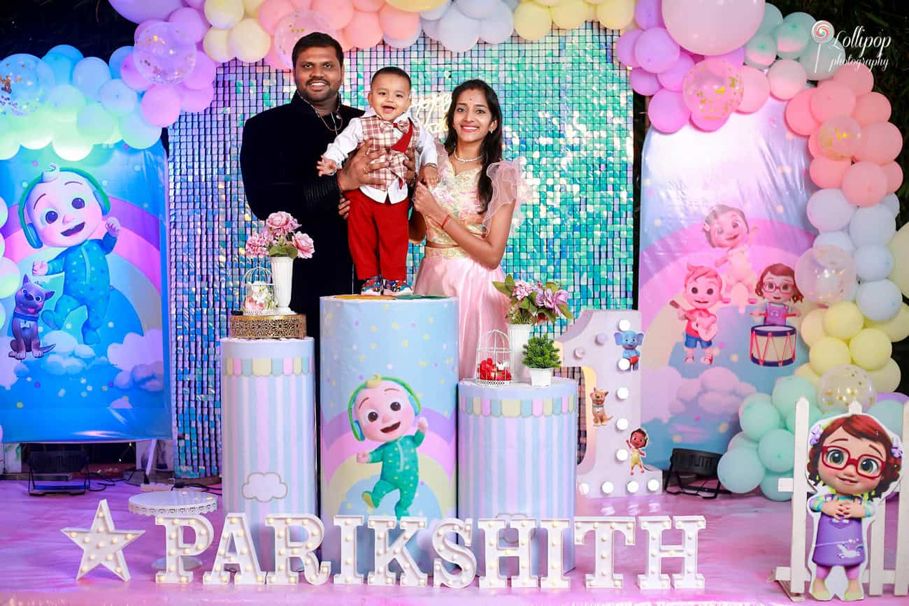 Family portrait at a birthday party with a colorful backdrop and pastel balloons, featuring a smiling toddler and his parents in Chennai, by Lollipop Photography.