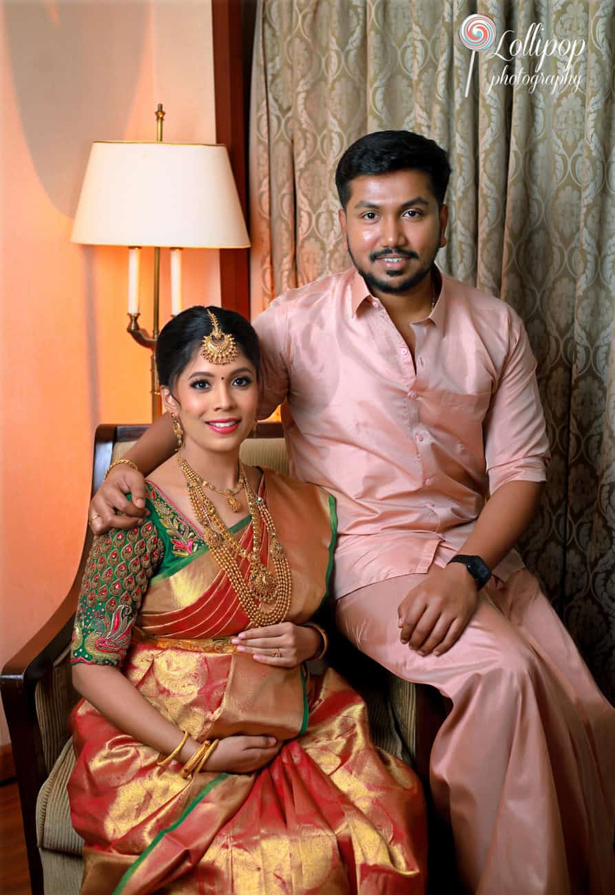 A beautiful expectant mother in a vibrant South Indian saree with her husband, both smiling warmly, seated comfortably in an indoor setting, highlighting a baby shower photography session in Chennai.