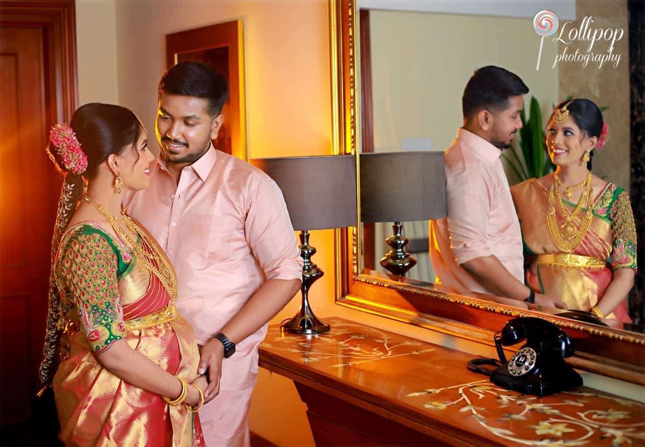 A couple smiles at each other, reflected in a large mirror, during a baby shower photo session; the pregnant woman is beautifully dressed in a traditional saree, enhancing the warmth of the indoor lighting in Chennai.