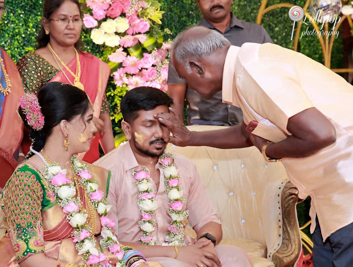 A traditional blessing ceremony with a family elder anointing the forehead of a radiant expectant mother, sitting beside her partner at their baby shower in Chennai.