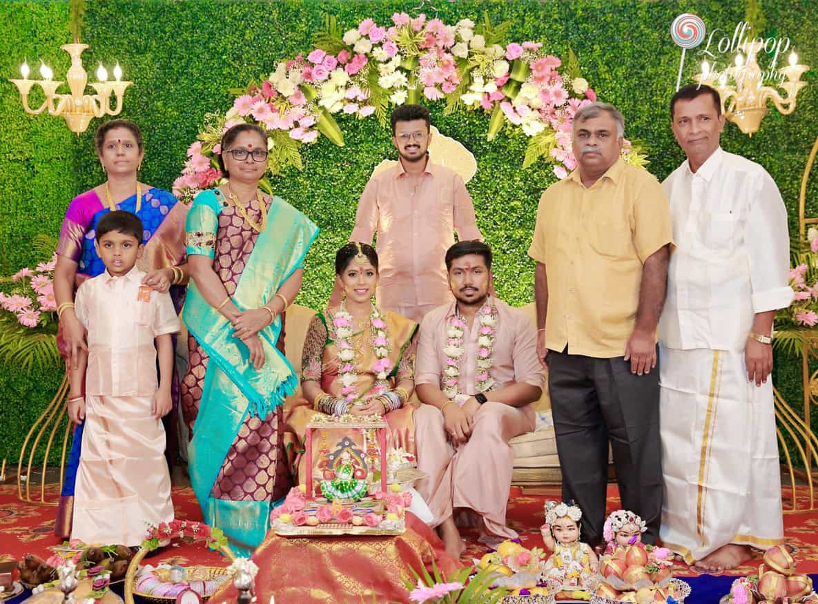 A vibrant group portrait at a baby shower, featuring the expectant couple seated in the middle with family members standing around them, against a backdrop of lush pink and yellow flowers in Chennai.