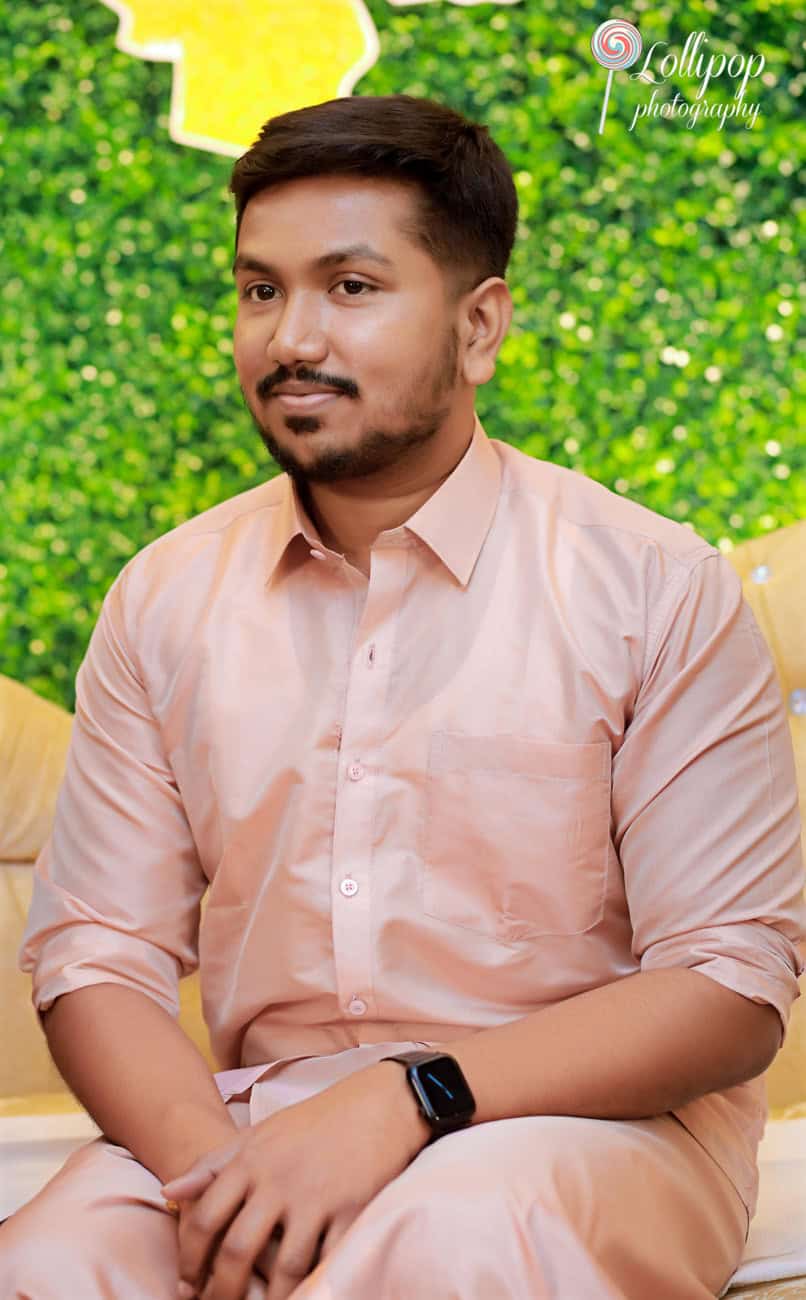 A portrait of a soon-to-be father wearing a peach-colored shirt, looking thoughtful and relaxed, set against a backdrop of lush greenery in Chennai.