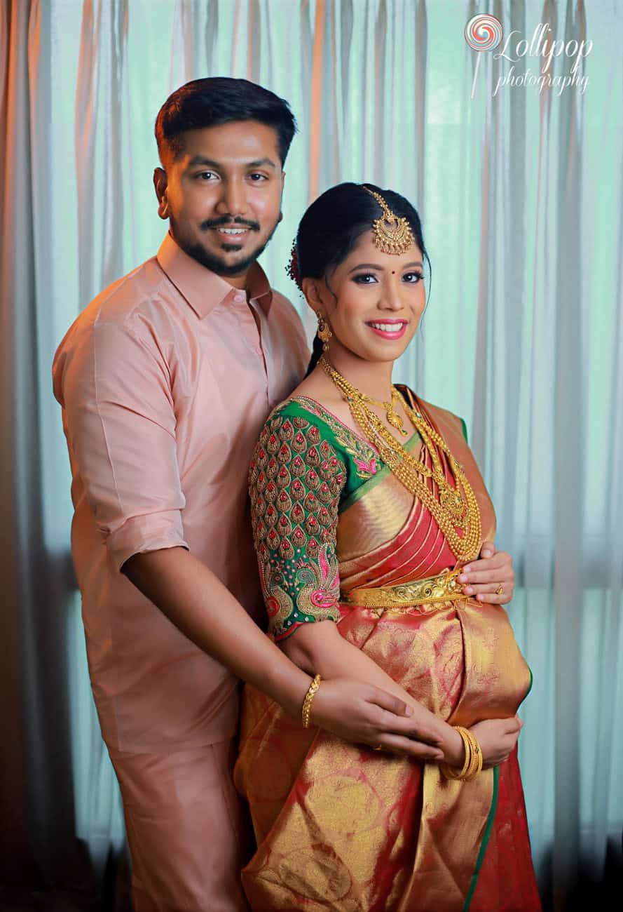 A glowing pregnant woman in a traditional green and red saree, embracing her partner, both sharing a joyful moment against a green curtain backdrop in Chennai.