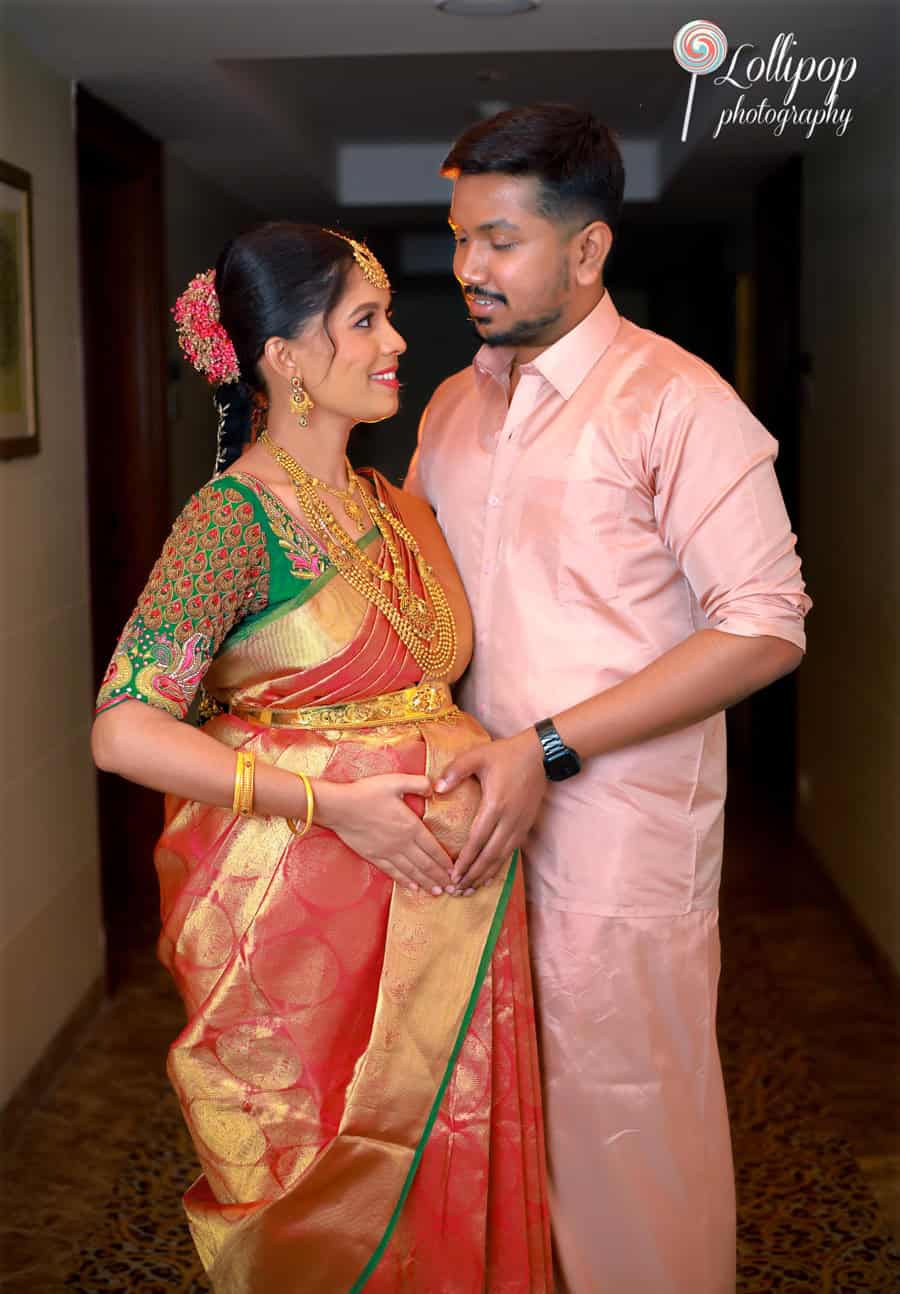 An intimate moment captured between an expectant couple, the woman adorned in a traditional saree and intricate gold jewelry, looking fondly at each other in a warmly lit hallway in Chennai.