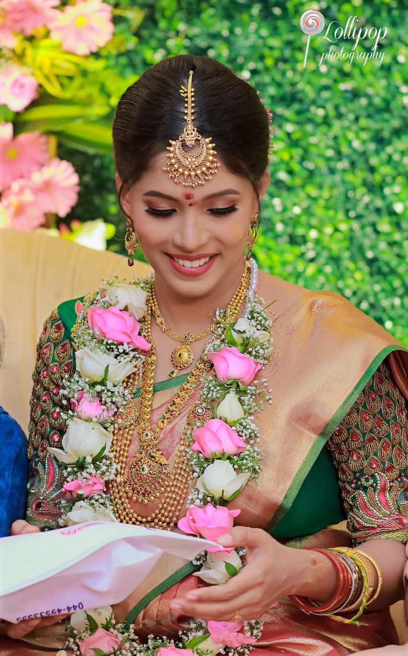 A glowing mother-to-be, dressed in traditional attire, happily reading heartfelt blessings during her baby shower, surrounded by a burst of pink and white floral decorations in Chennai.