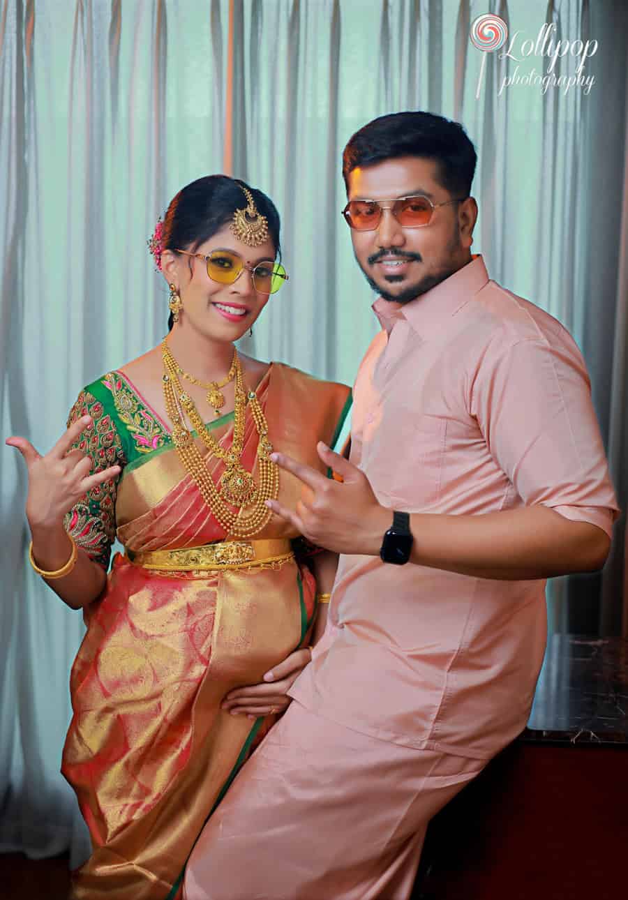 A playful moment with a pregnant couple, the woman in a traditional saree adorned with gold jewelry, giving a fun pose with her partner against a green curtain backdrop in Chennai.