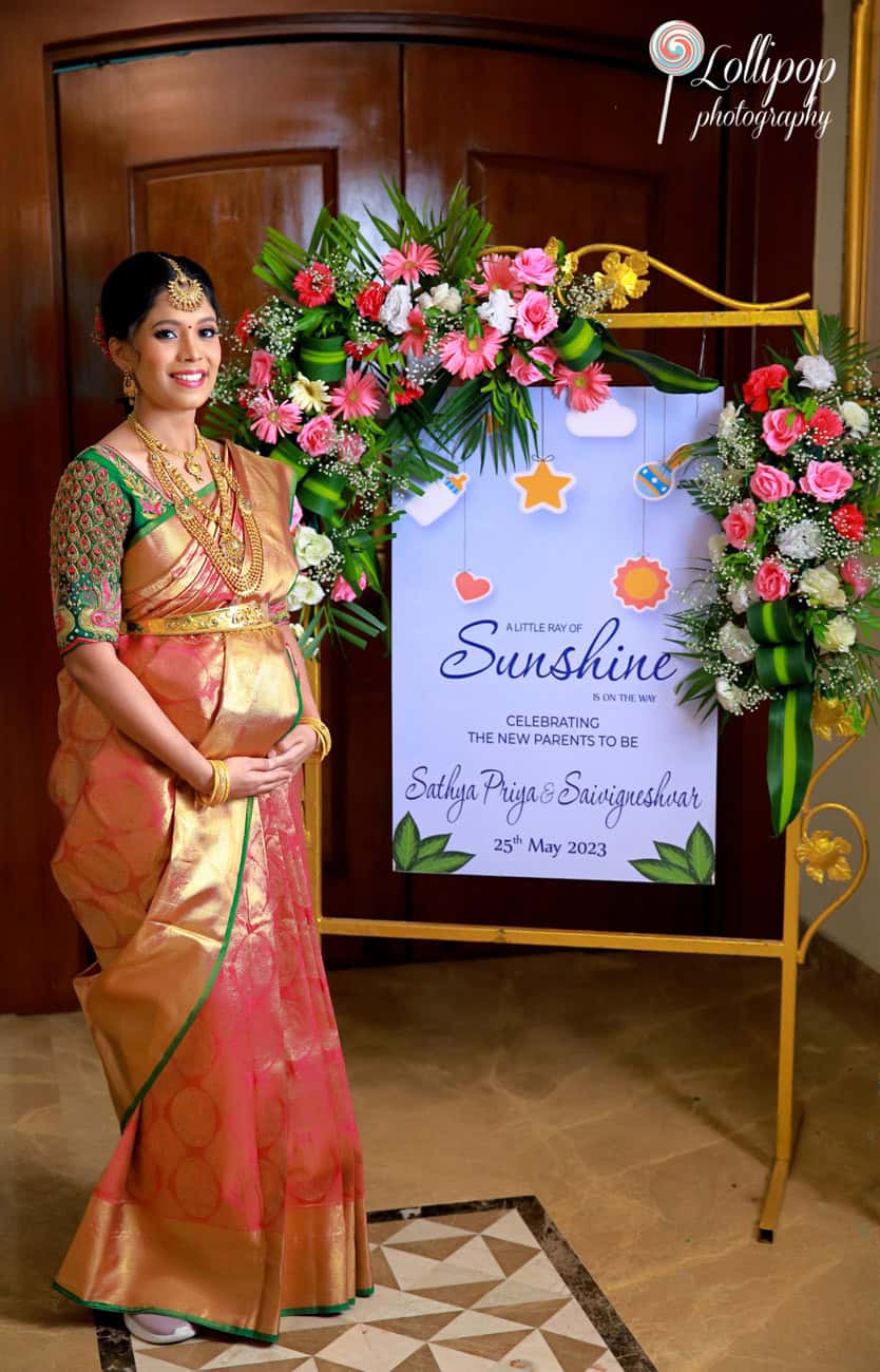 A radiant pregnant woman in traditional attire stands beside a festive floral arrangement at a baby shower event, her joyful expression matching the colorful decor in Chennai.