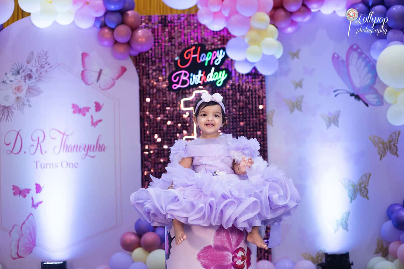 Lavender-themed birthday celebration for a joyful toddler at her first birthday party, with colorful balloons and a bright neon 'Happy Birthday' sign, captured by Lollipop Photography in Chennai.