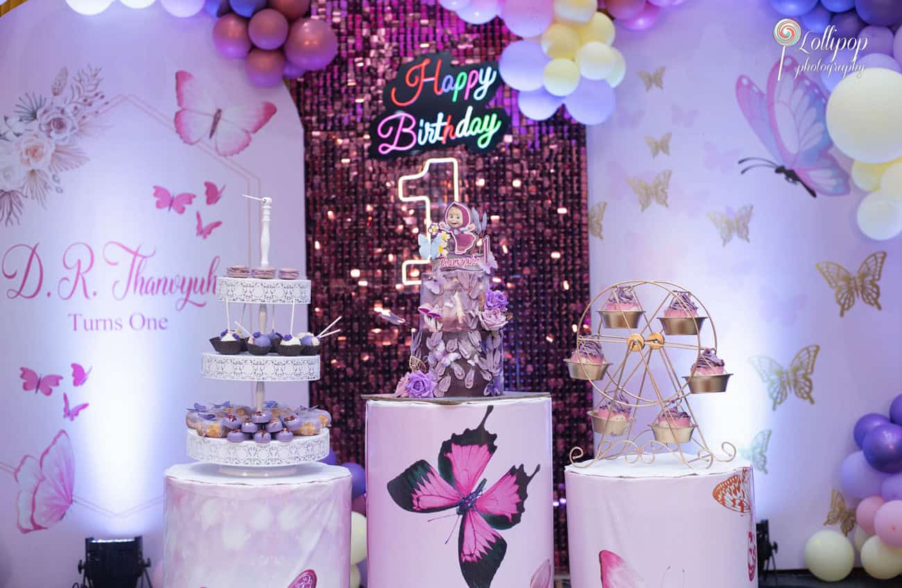 Exquisite birthday cake display featuring multiple layers and a custom topper at a first birthday party in Chennai, with vibrant balloons and butterfly decorations by Lollipop Photography