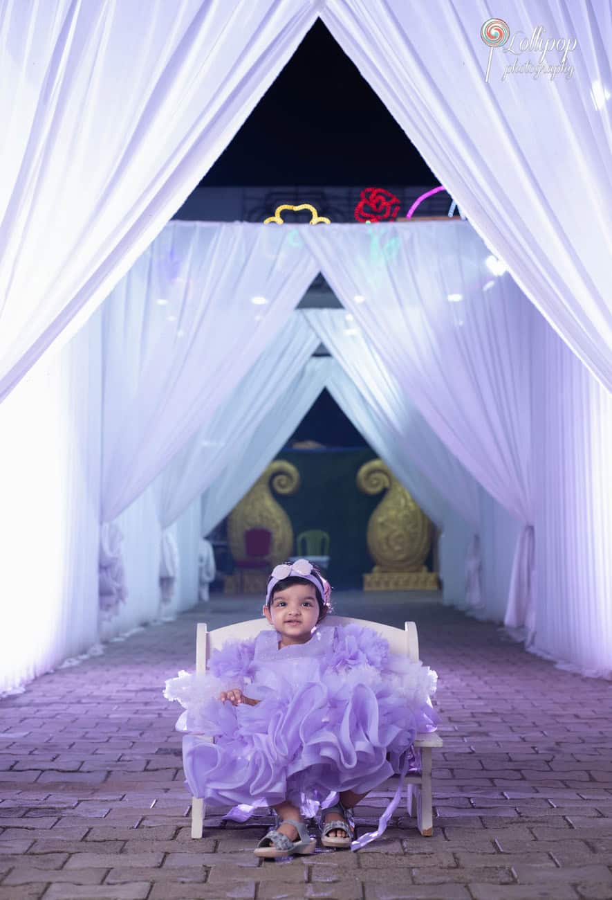 Little birthday girl sitting on a miniature throne under an elegantly draped white archway, wearing a lavender dress at her birthday celebration in Chennai, beautifully captured by Lollipop Photography