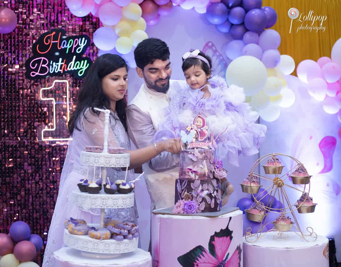 Parents assisting their daughter in cutting her first birthday cake at a lavishly decorated party in Chennai, with a butterfly theme, by Lollipop Photography