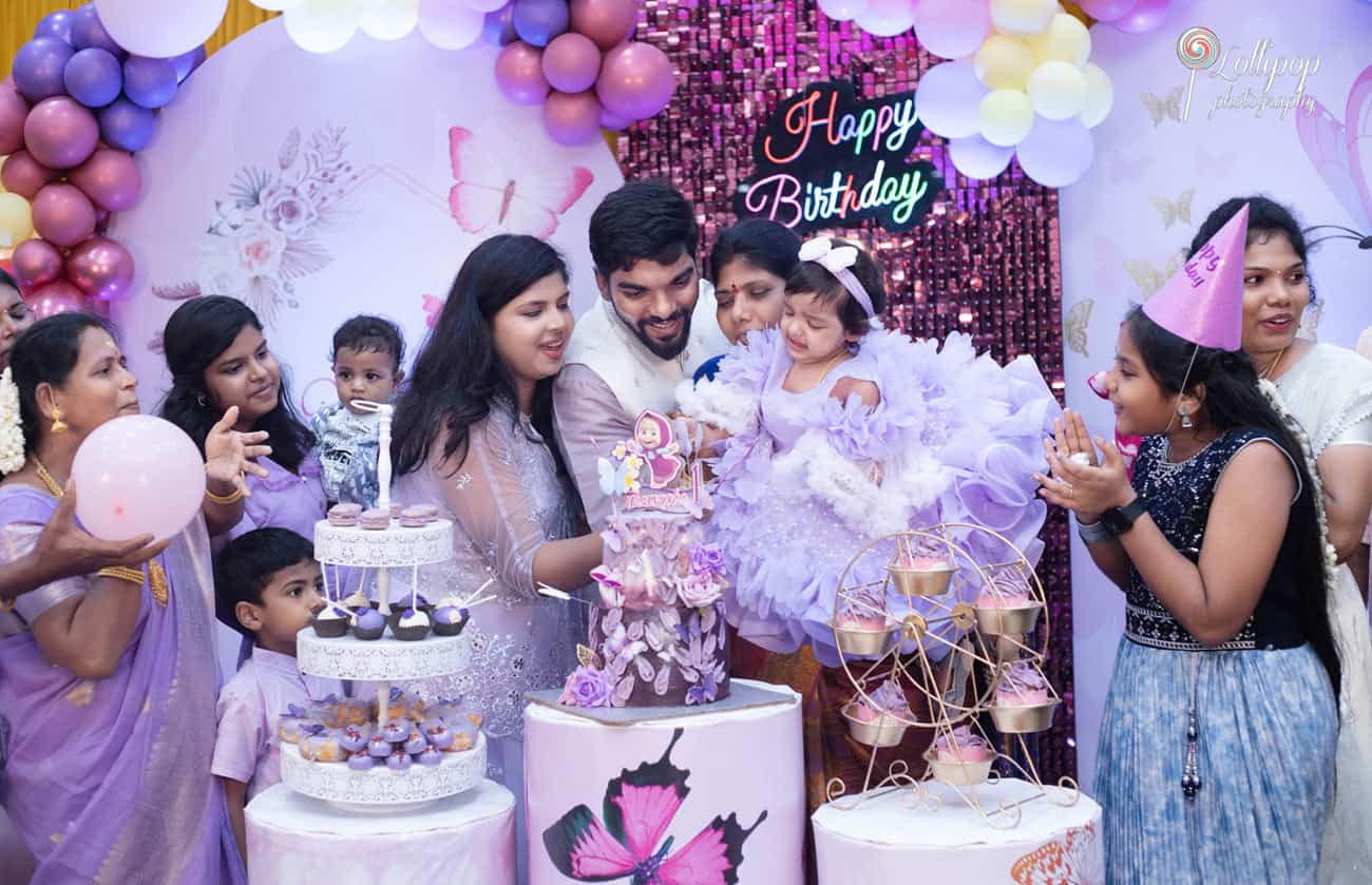 Family and friends gathered around the birthday cake as the little girl reaches for the topper, a vibrant scene full of smiles and joy at a first birthday party in Chennai, captured by Lollipop Photography.