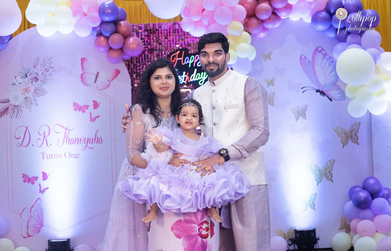 Happy family of three celebrating the first birthday of their daughter, Thanvyuha, standing in front of a butterfly-themed backdrop adorned with balloons