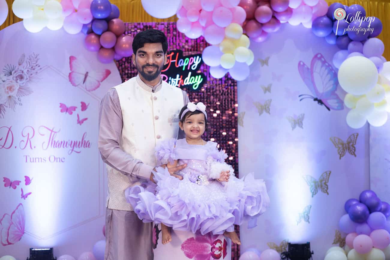 Father smiling as he holds his daughter, who is dressed in a festive lavender birthday outfit, celebrating her first birthday in Chennai.