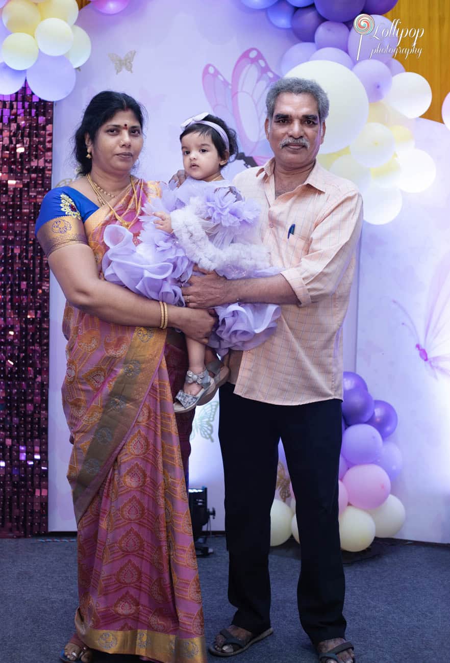 grandparents lovingly holding their great-daughter on her first birthday, adorned in traditional Indian attire and a lavender dress, photographed by Lollipop Photography in Chennai.