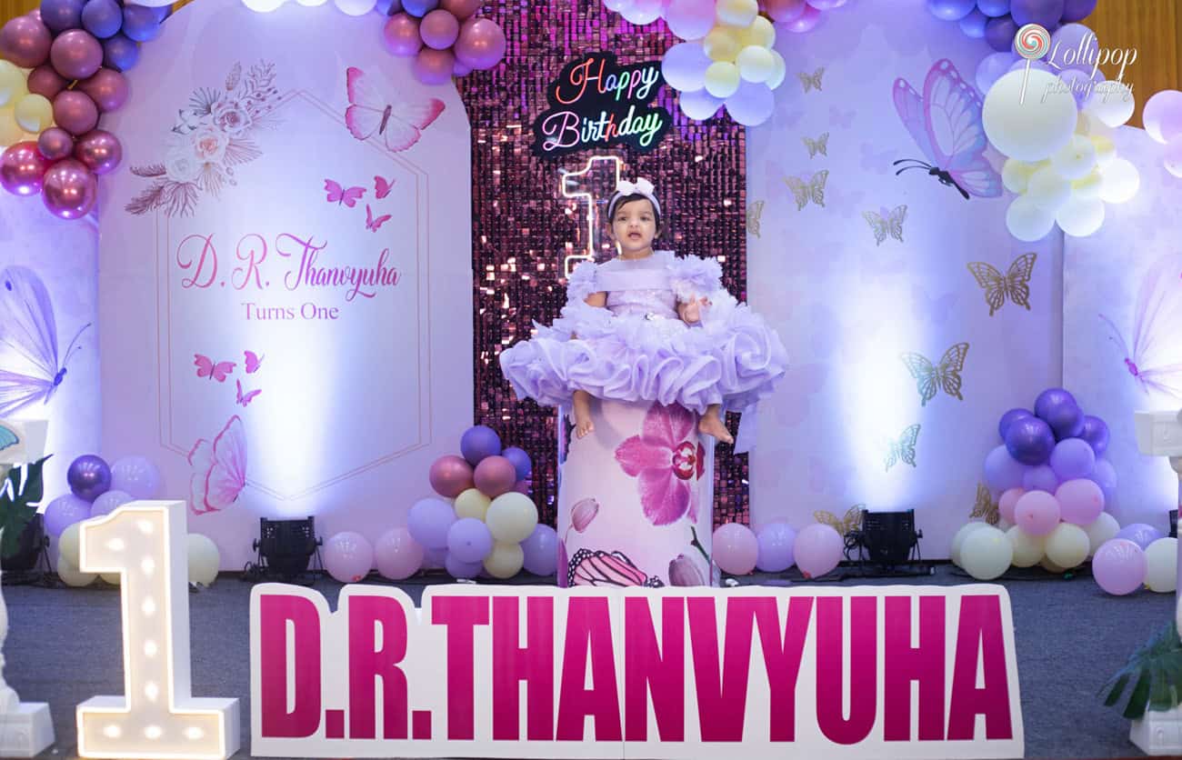 Elegant first birthday party in Chennai featuring a toddler in a lush lavender dress, surrounded by balloons and a custom birthday backdrop, photographed by Lollipop Photography