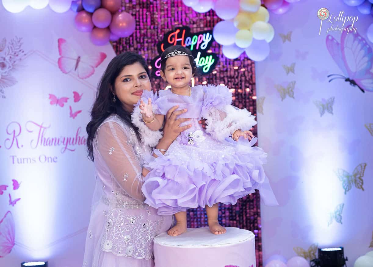 Mother tenderly holding her daughter in a lavish lavender birthday outfit at her first birthday party, adorned with balloons and butterfly decorations, captured by Lollipop Photography in Chennai.