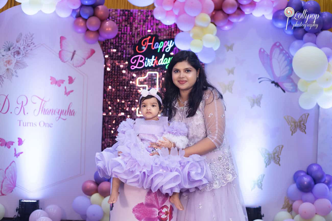 Mother holding her daughter dressed in a fluffy lavender dress at her first birthday celebration, with a colorful balloon backdrop.