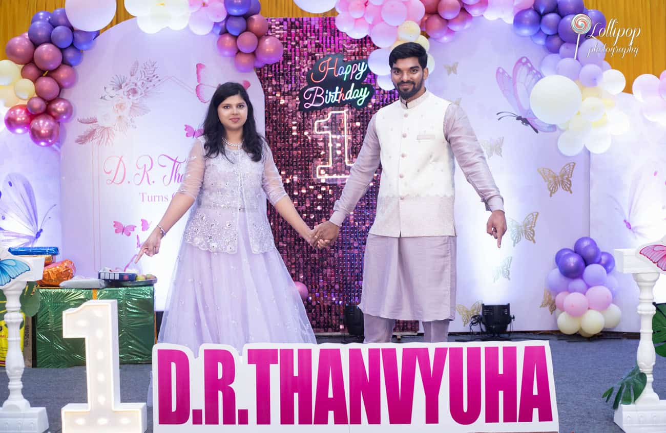 Parents holding hands during their daughter's first birthday party in Chennai, creating a heartwarming scene under a butterfly-themed backdrop, captured by Lollipop Photography