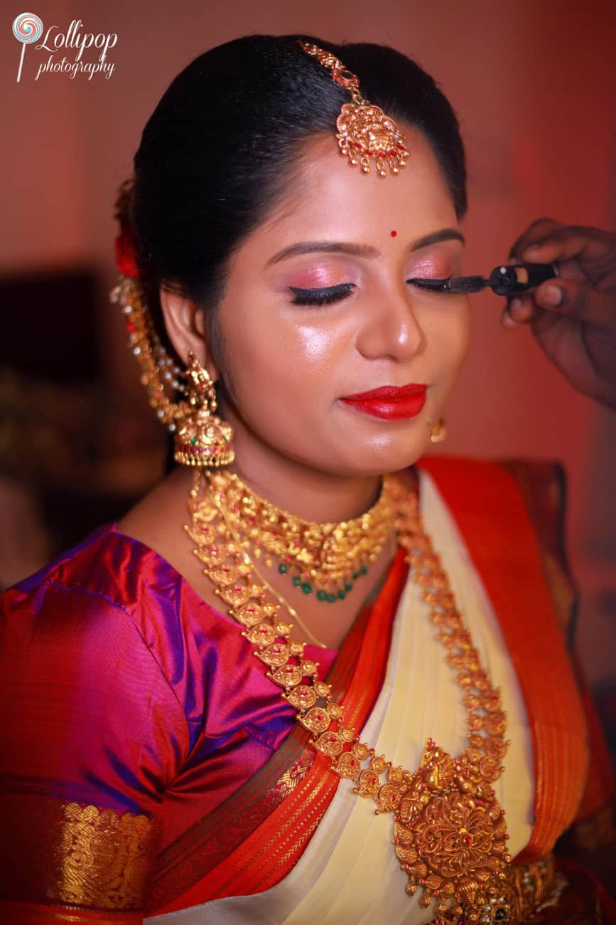 Close-up of a makeup artist applying mascara to an expectant mother adorned in traditional bridal jewelry, capturing the preparation moments of a baby shower in chennai, by Lollipop Photography.