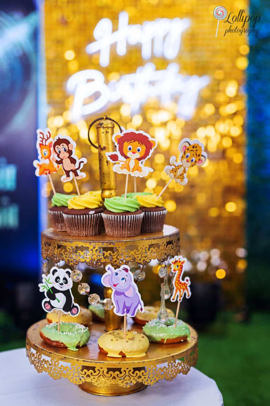 Vibrantly decorated cupcakes with jungle animal toppers set against a golden 'Happy Birthday' backdrop, creating a festive scene at a first birthday party, photographed by Lollipop Photography in Chennai.