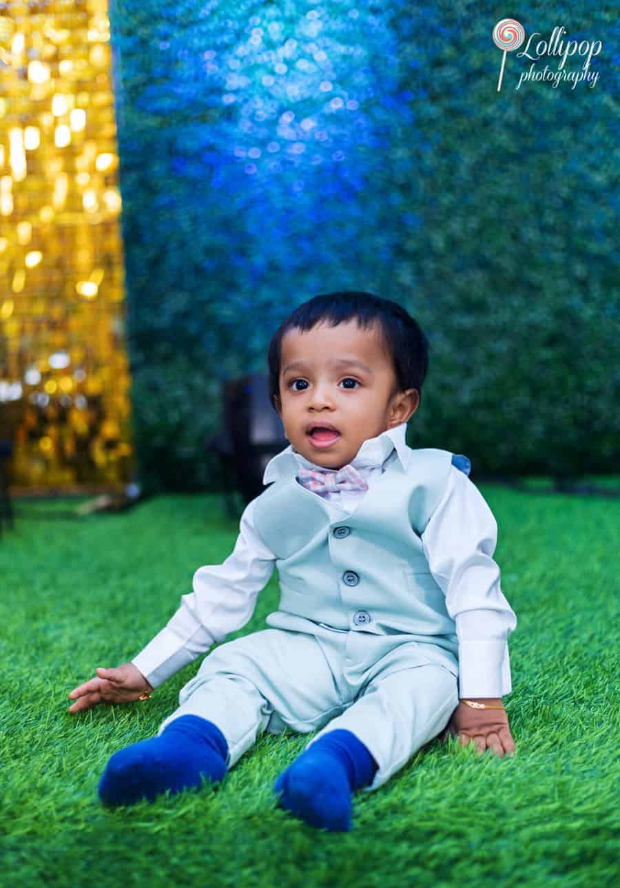 Adorable baby boy dressed in a smart white outfit sitting on lush green grass, with a vibrant blue and gold backdrop, during his first birthday photoshoot by Lollipop Photography in Chennai