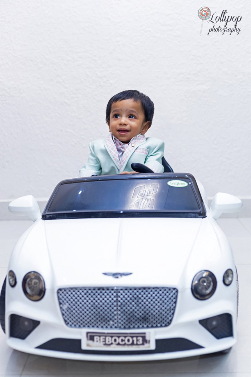 Adorable baby boy smiling while seated in a miniature white Bentley, making a memorable moment at his first birthday party, photographed by Lollipop Photography in Chennai