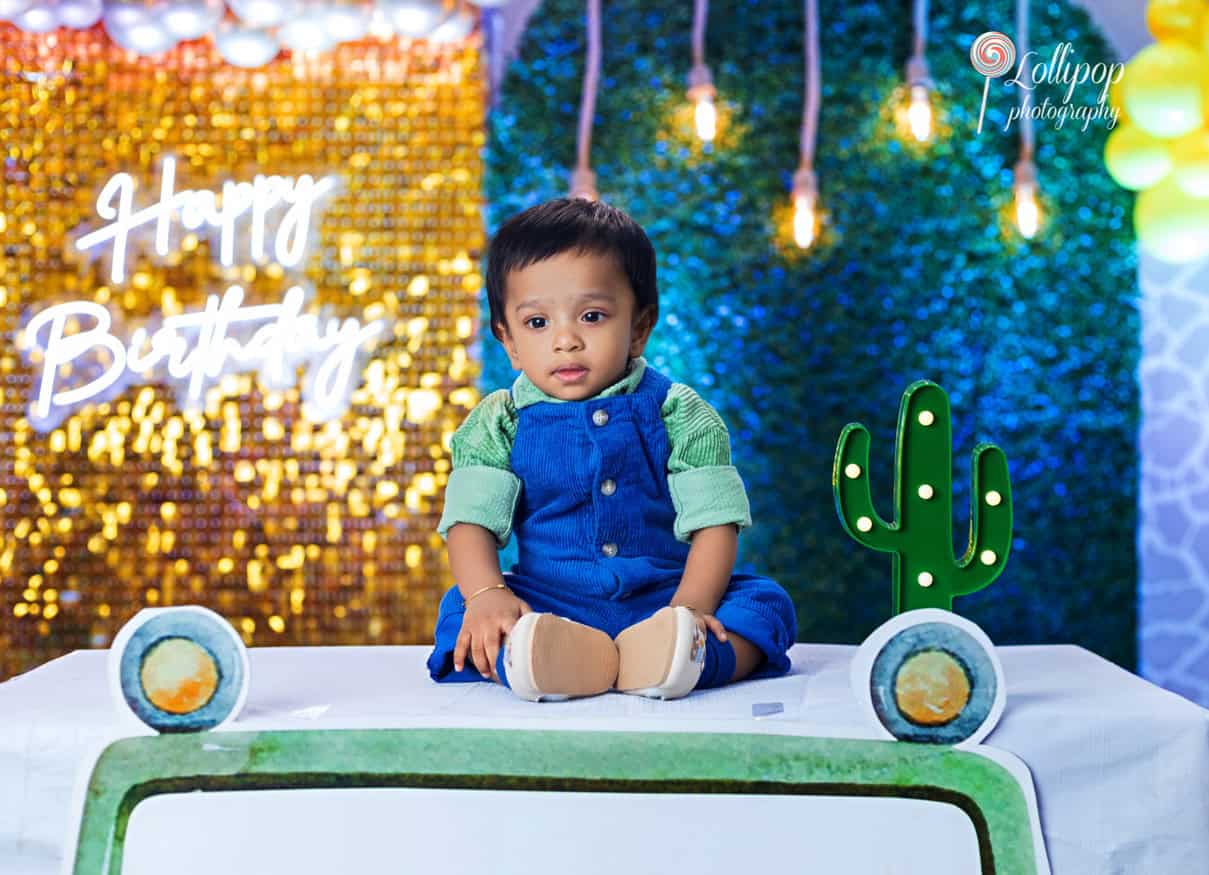 Cute baby boy seated on a creatively decorated vehicle with cactus decoration and a vibrant 'Happy Birthday' backdrop, photographed by Lollipop Photography in Chennai.