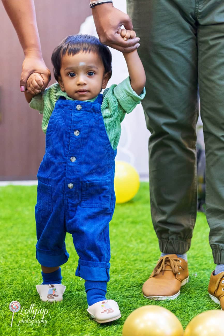 First steps captured beautifully at a first birthday celebration, with a baby in blue overalls held up by parents, surrounded by gold balloons, by Lollipop Photography in Chennai