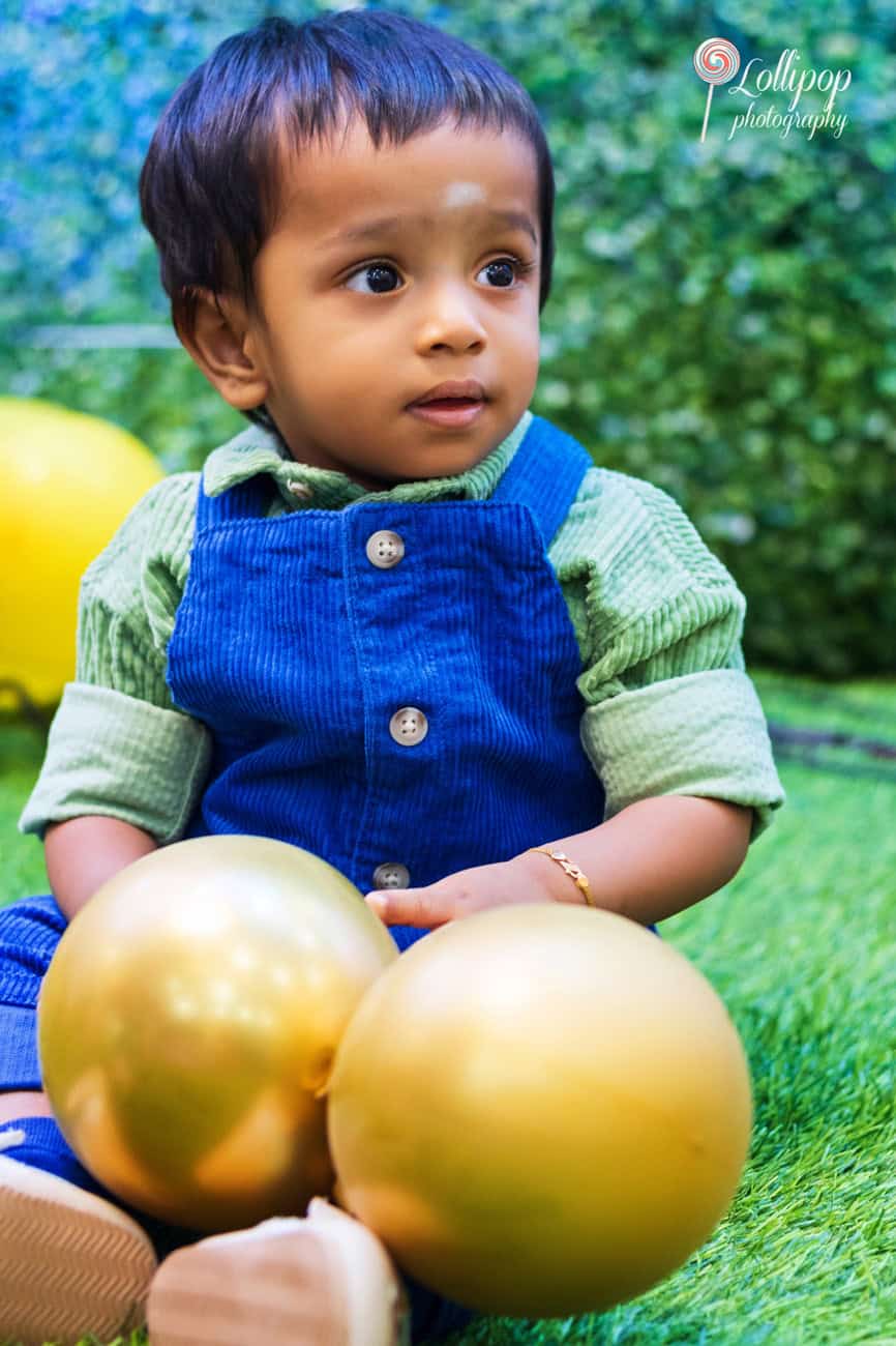 Adorable baby sitting among golden balloons on a lush green lawn, looking curious and cute, during a first birthday photoshoot with Lollipop Photography in Chennai