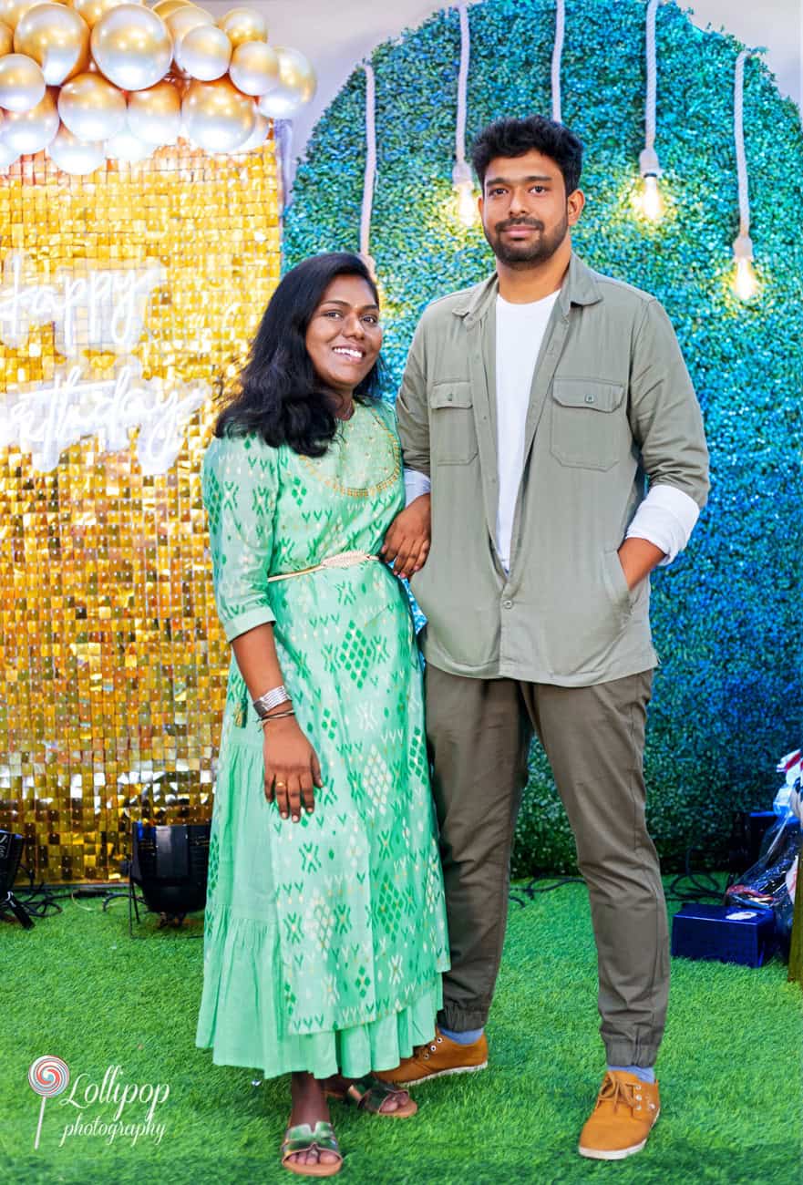 Loving couple standing together with a radiant smile in front of a dazzling birthday backdrop of golden balloons and greenery, captured by Lollipop Photography in Chennai