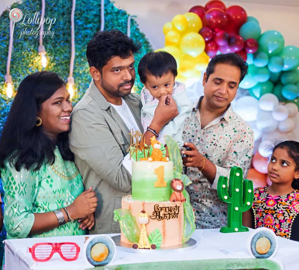 Extended family enjoying a vibrant first birthday party with a colorful jungle-themed cake, commemorated by Lollipop Photography in Chennai.