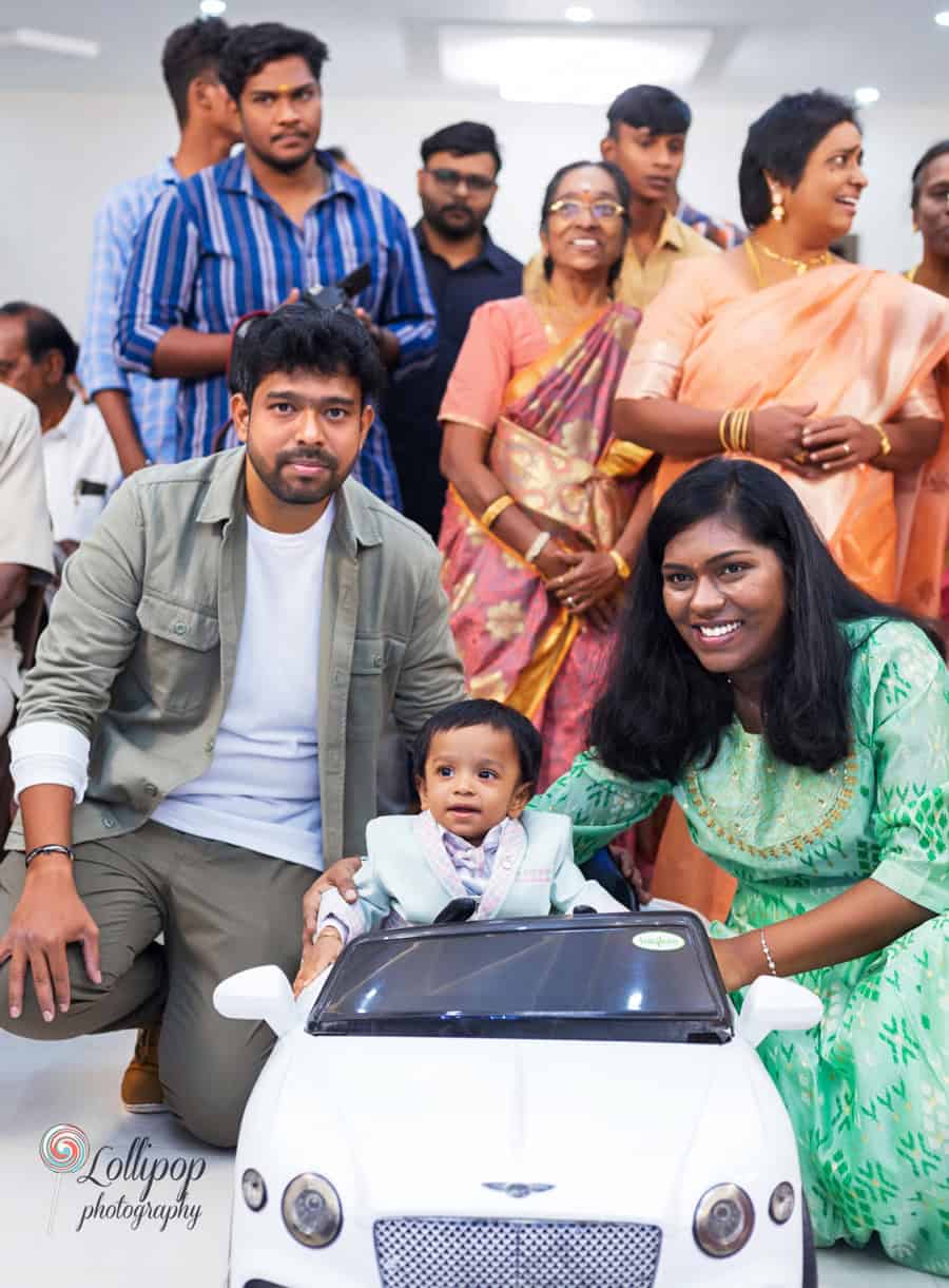 Family and guests smiling and gathering around a baby boy in a toy car during his first birthday celebration, captured by Lollipop Photography in Chennai