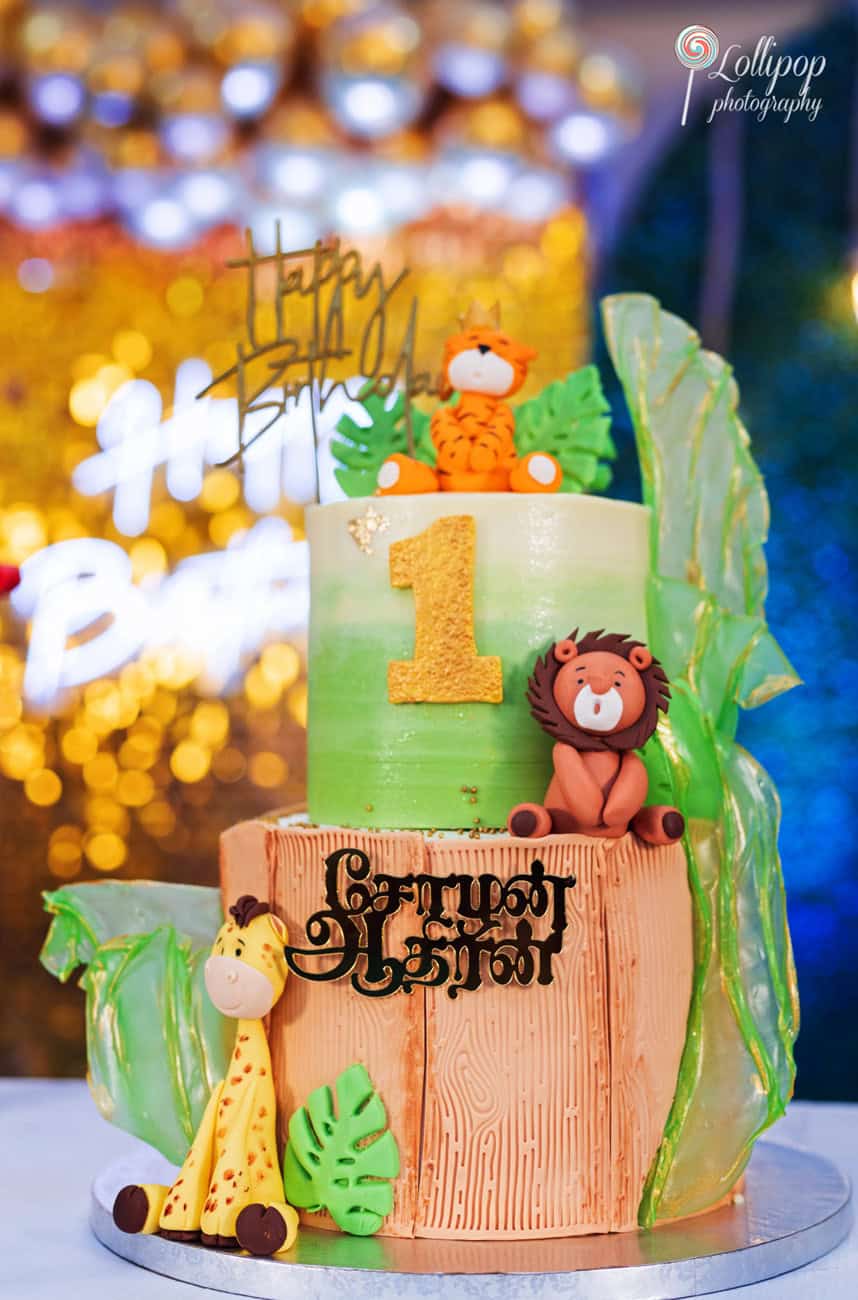 Exquisite first birthday cake with a jungle theme featuring fondant animals and a glittering 'Happy 1st Birthday' topper, skillfully captured by Lollipop Photography in Chennai.