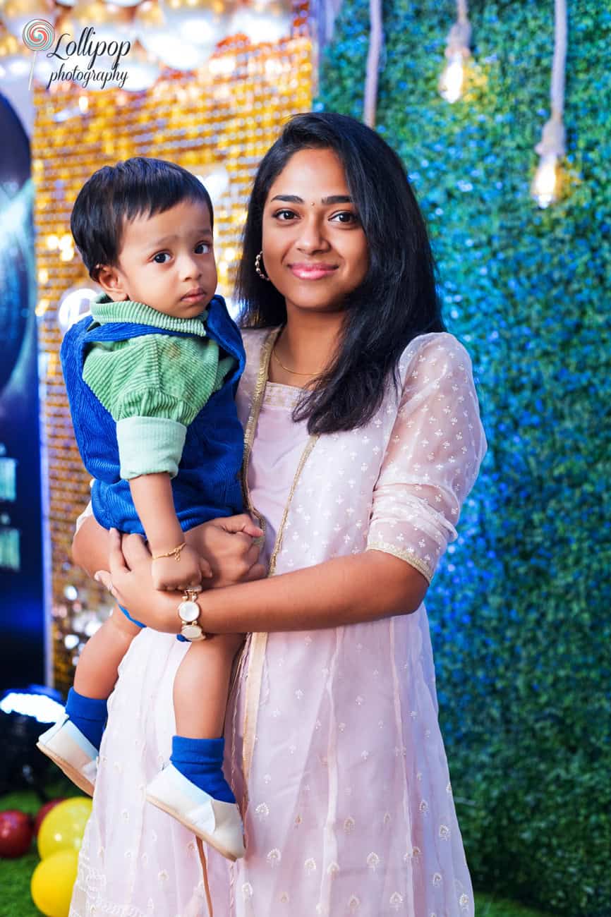 Mother in a beautiful pink outfit holding her baby, with a festive gold and blue backdrop during a birthday photoshoot by Lollipop Photography in Chennai