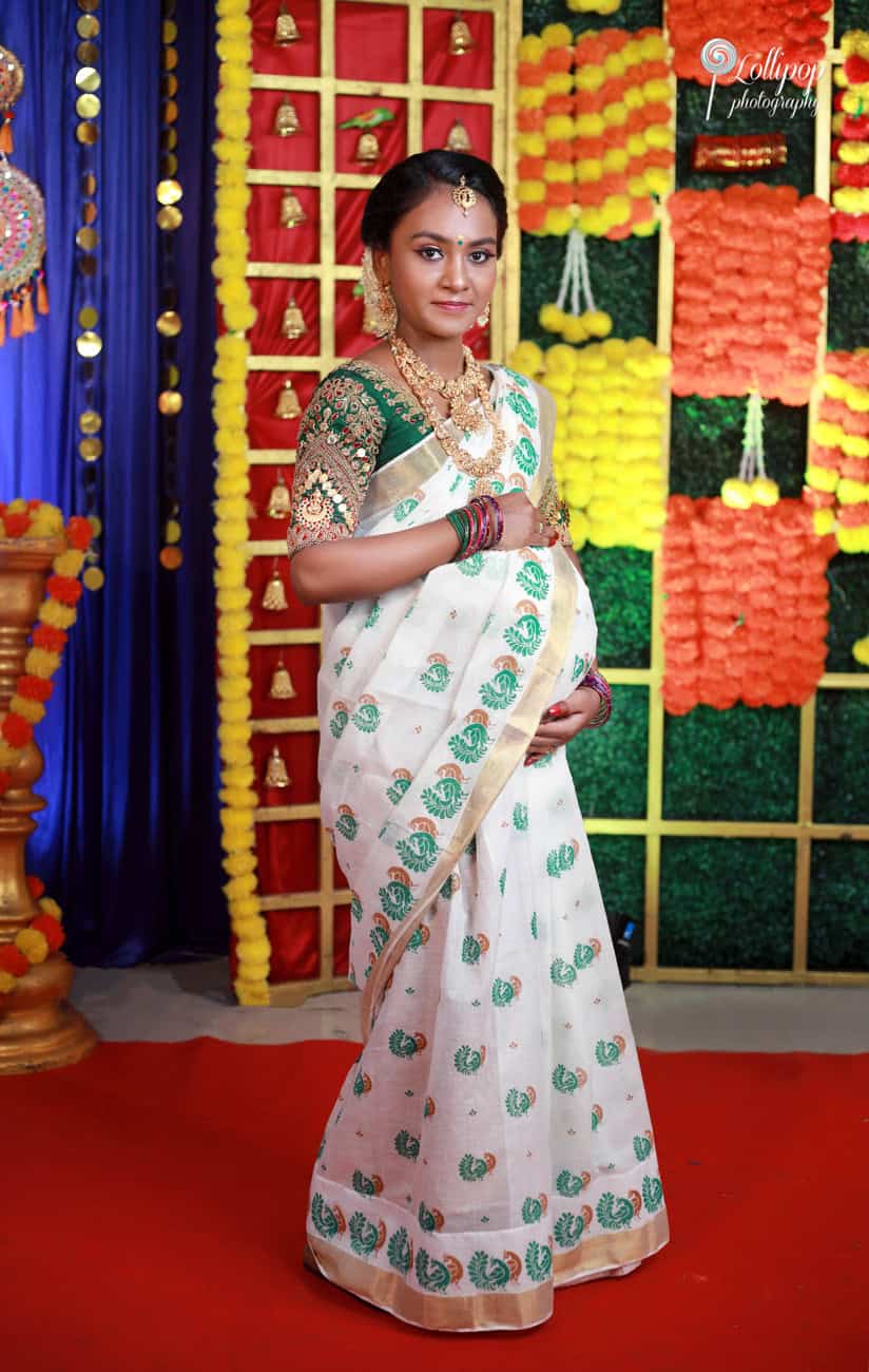 Expectant mother in a traditional white and green saree adorned with gold jewelry, posing at a beautifully decorated venue with vibrant floral and bell decorations, Lollipop Photography, Coimbatore