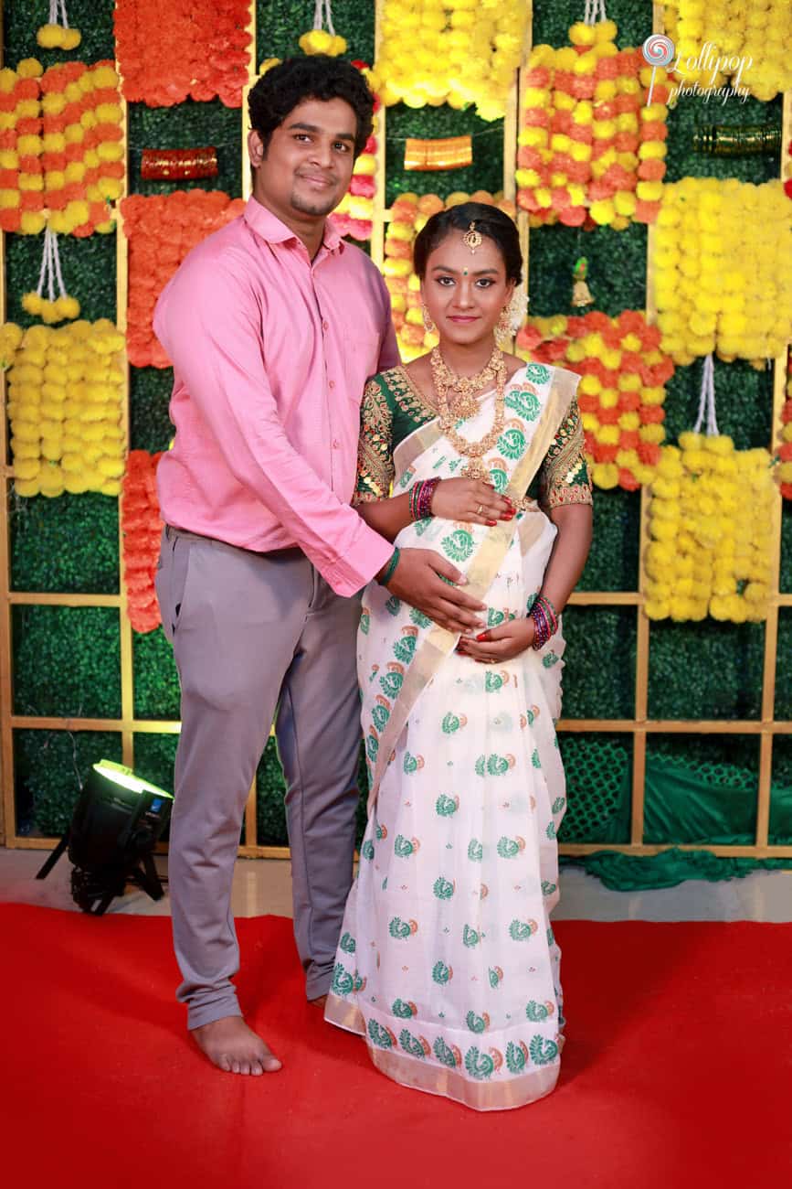 Maternity photoshoot of a couple in traditional attire, standing together with a vividly decorated backdrop of flowers and bells, capturing their joyful anticipation in Coimbatore