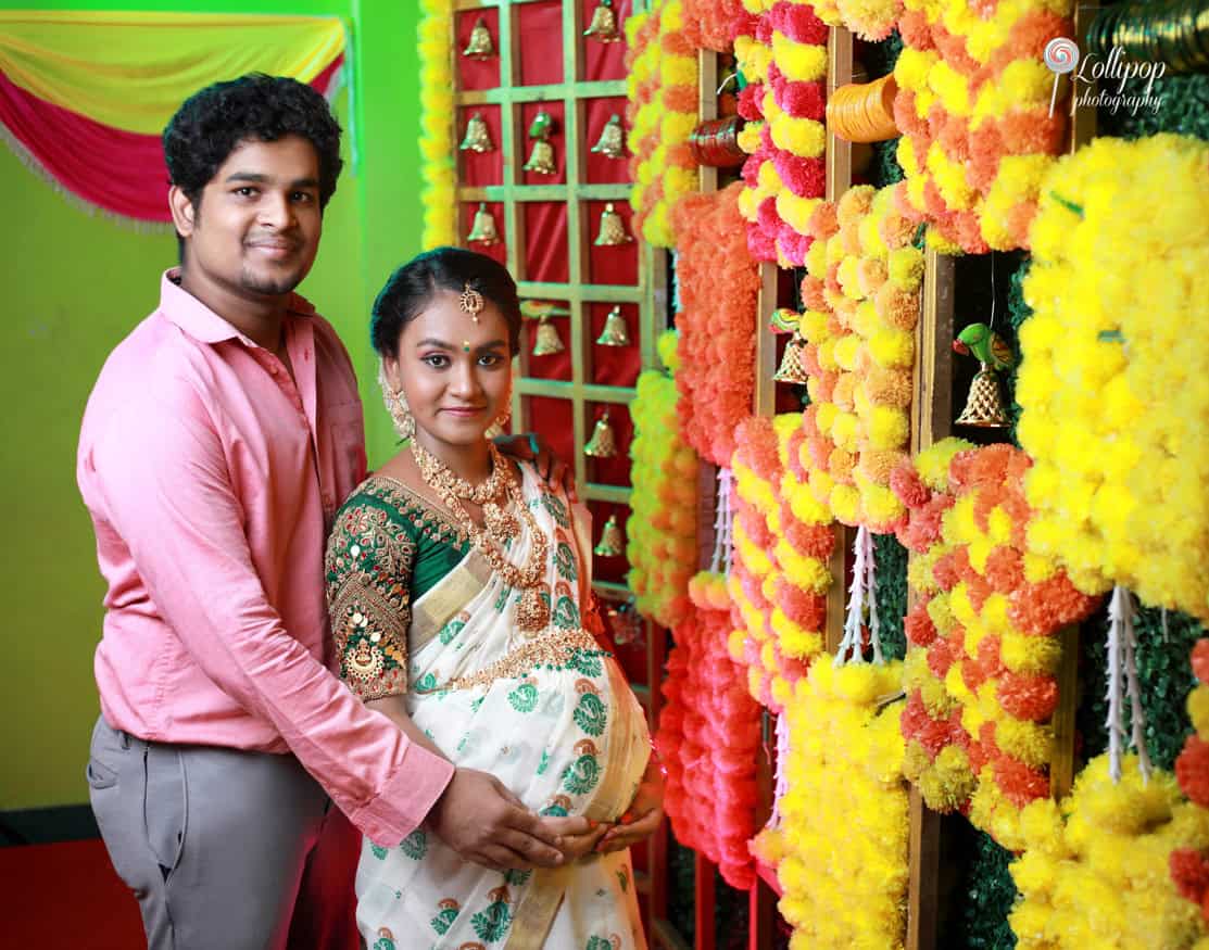 Couple photo session at a maternity event, with the expectant mother in a traditional saree and her partner in a pink shirt, both smiling beside a colorful, culturally themed backdrop in Coimbatore.