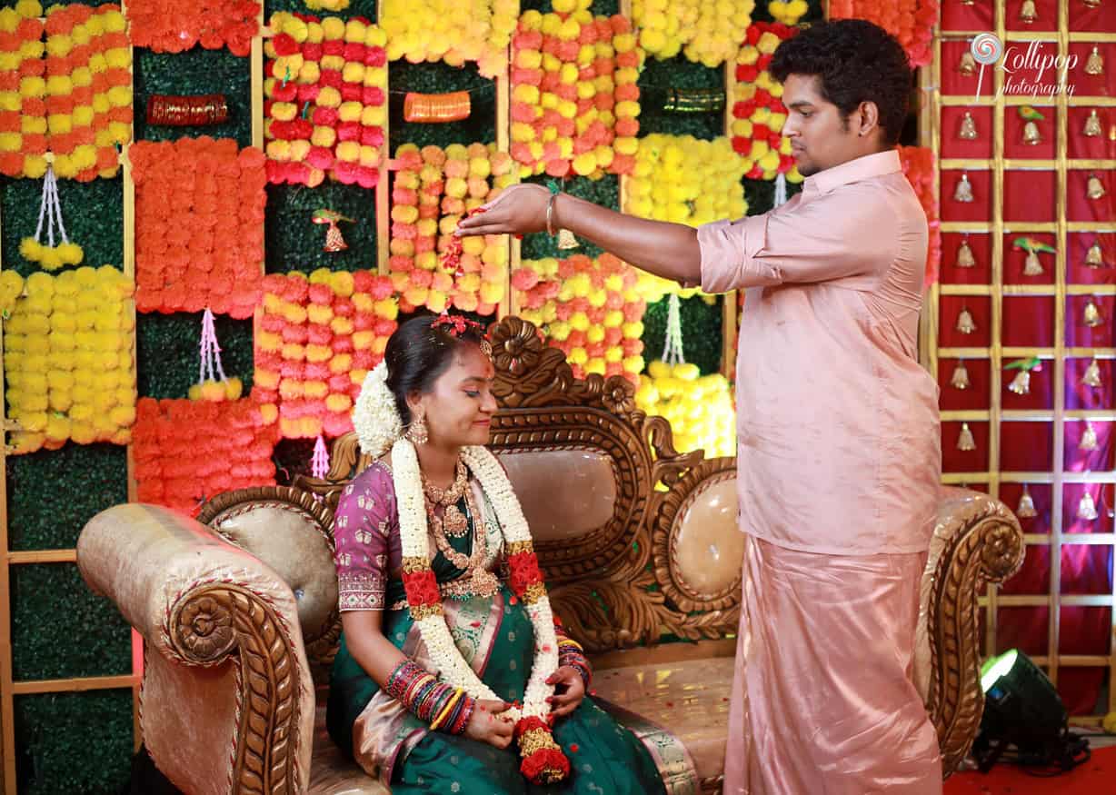 Playful maternity moment with a husband playfully blessing his seated wife, creating a lively atmosphere amidst colorful baby shower decorations in Coimbatore