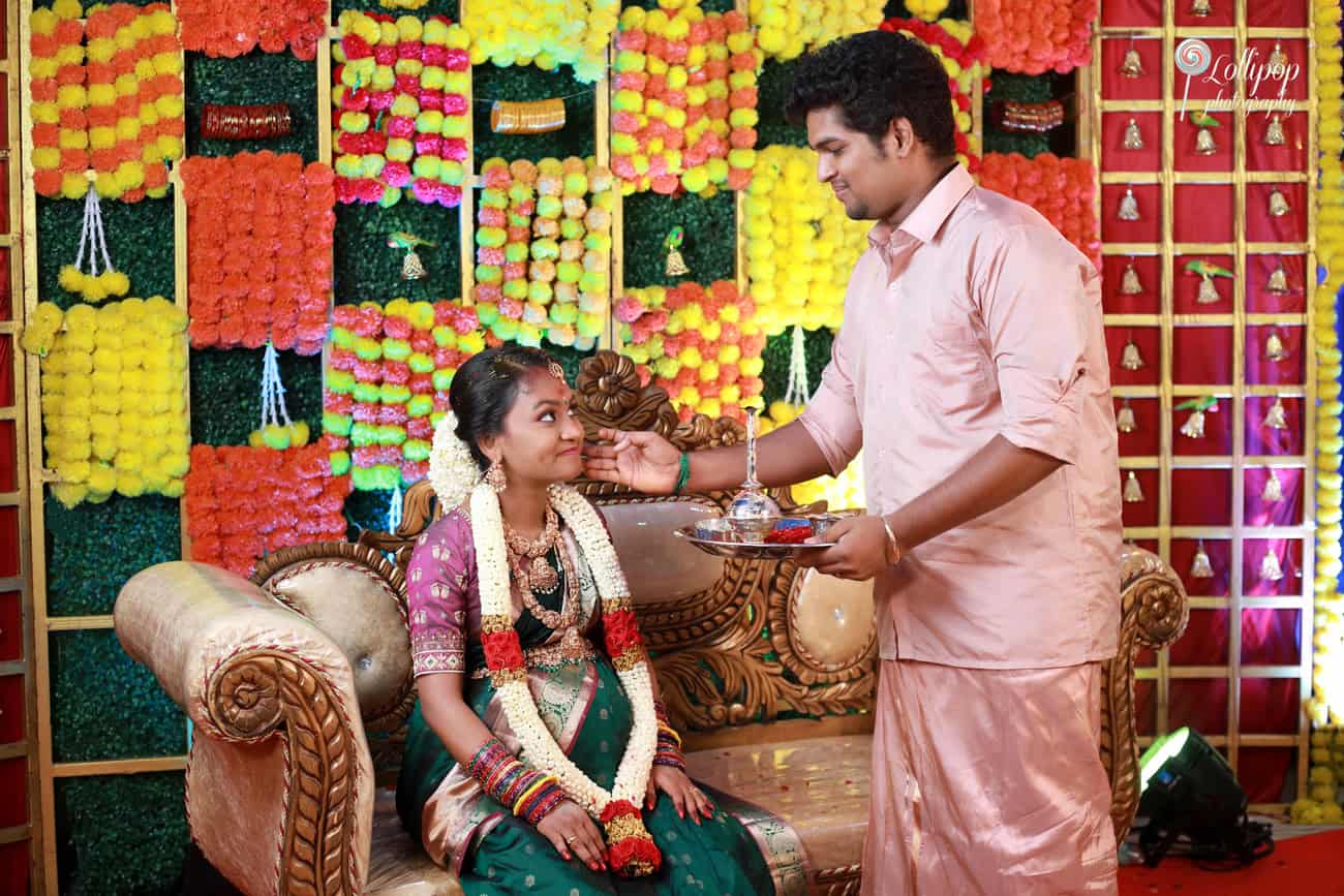 Tender moment as a husband applies sandalwood paste on his wife's forehead during their maternity celebration, surrounded by rich cultural decor in Coimbatore