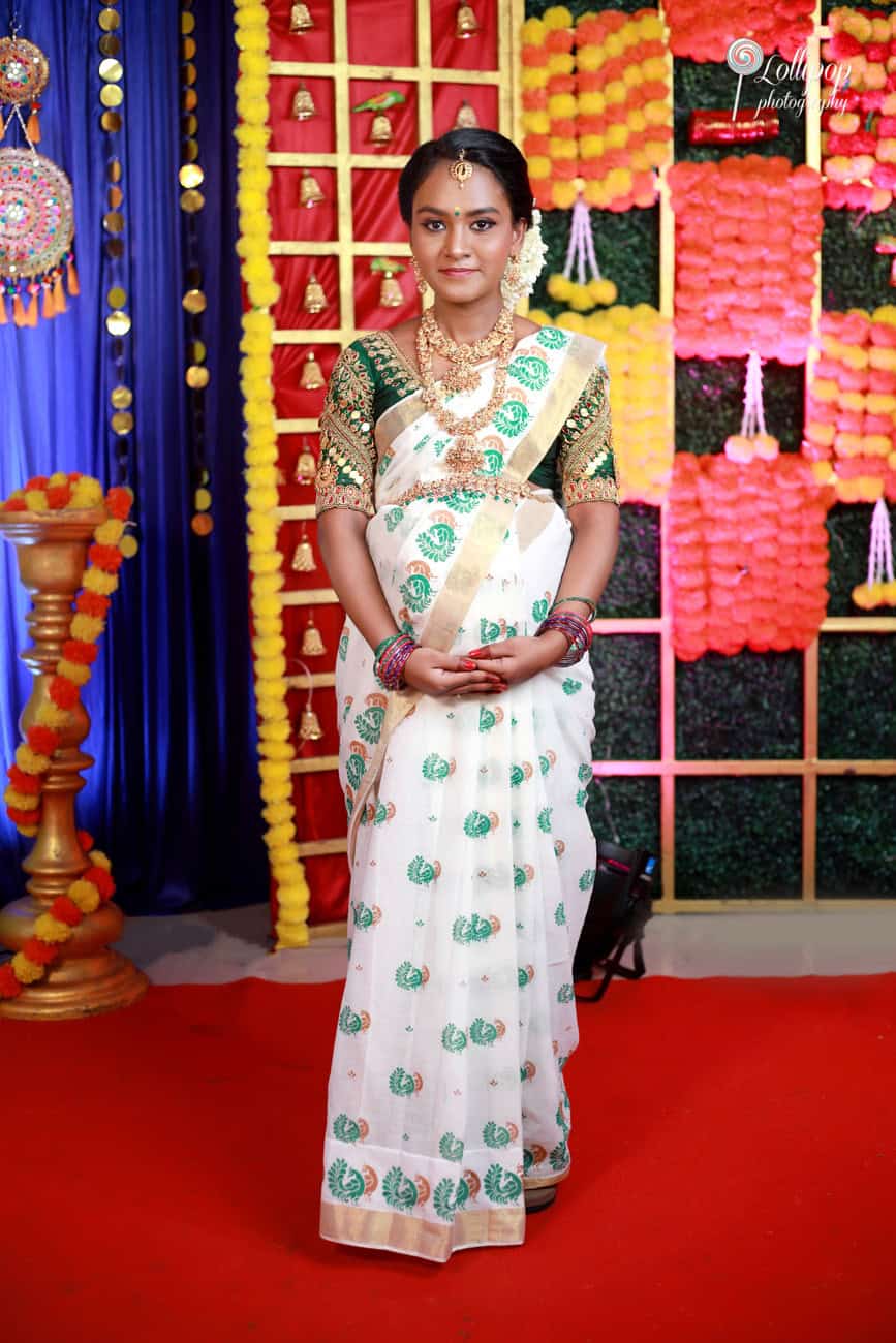 Pregnant woman dressed in a traditional saree with intricate embroidery and jewelry, standing against a backdrop of colorful flower garlands and cultural decorations, Coimbatore.