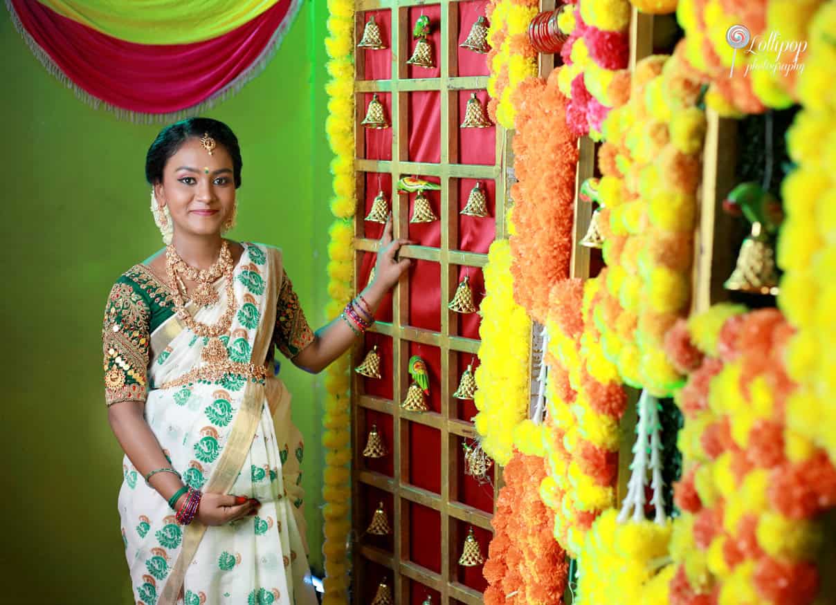 Expectant mother in traditional attire smiling beside a decorative wall with golden bells and multicolored garlands, enhancing the festive atmosphere of her maternity photoshoot in Coimbatore.