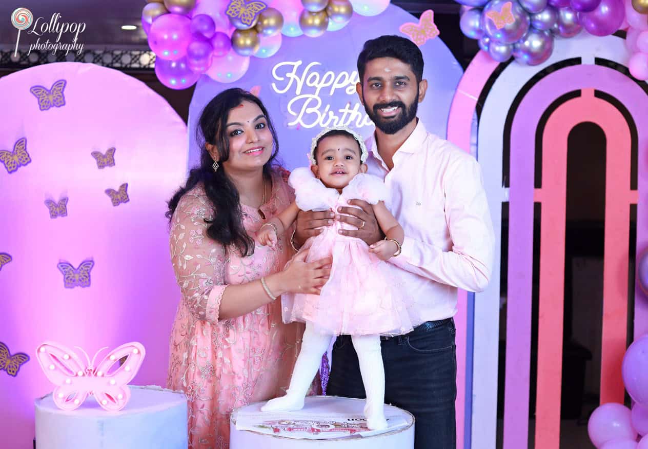 Smiling family of three posing in front of a colorful birthday setup with balloons and butterfly decorations, captured by Lollipop Photography in Chennai.