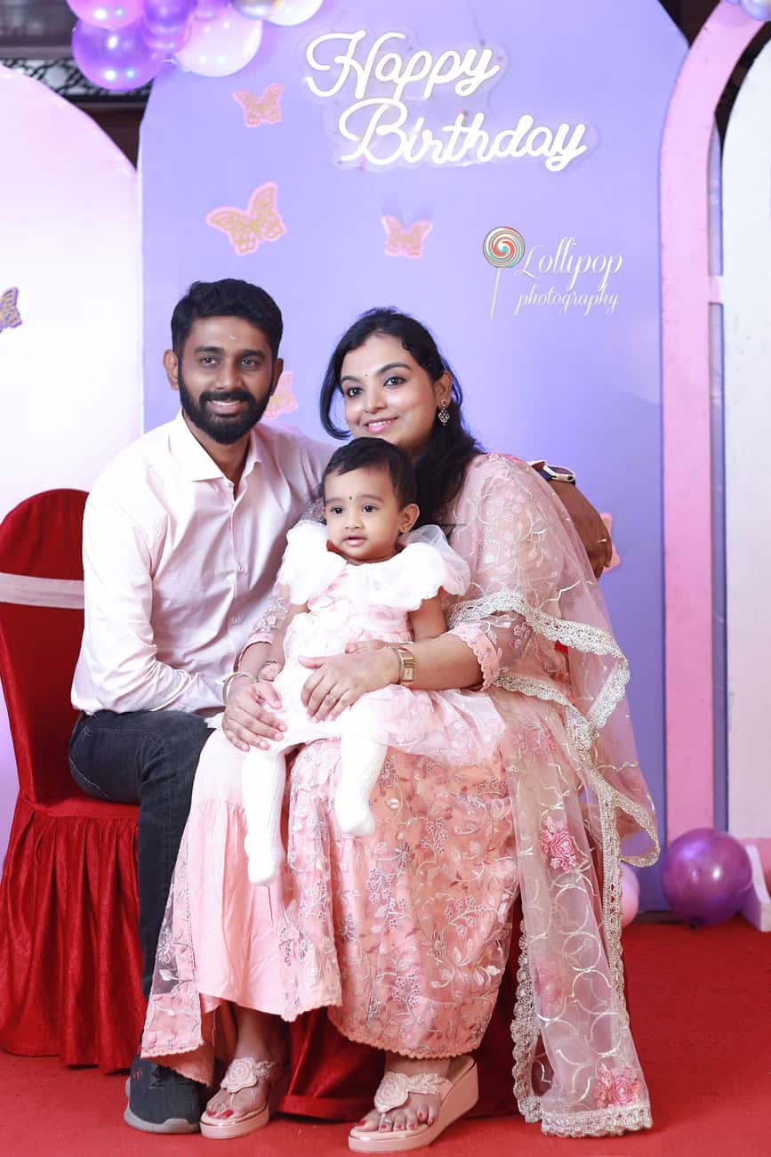 A happy family poses with their baby in front of a festive birthday backdrop featuring balloons and butterflies, captured in Chennai by Lollipop Photography.