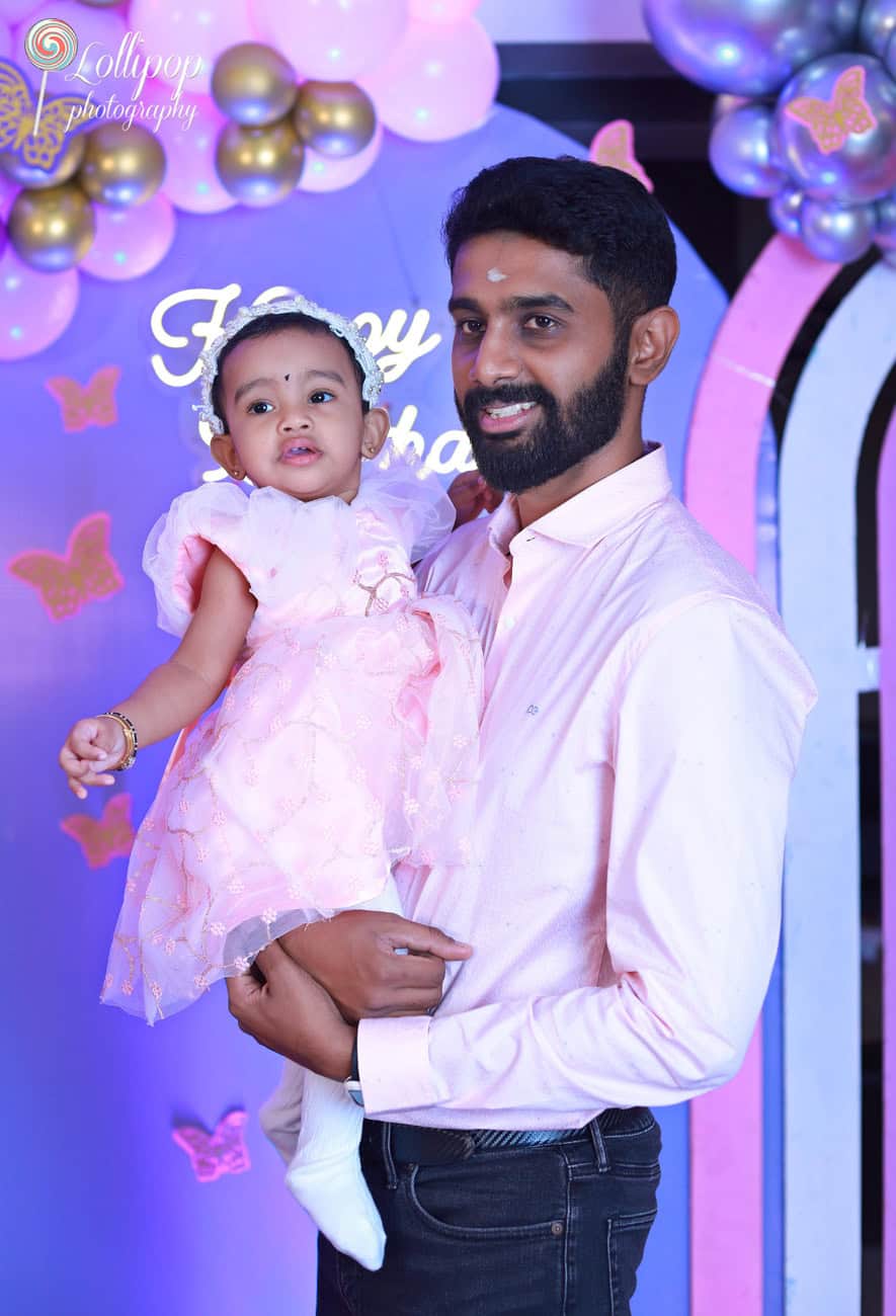 A father holds his little daughter dressed in a festive pink outfit during her first birthday celebration, with a whimsical backdrop of balloons and butterflies, by Lollipop Photography in Chennai.