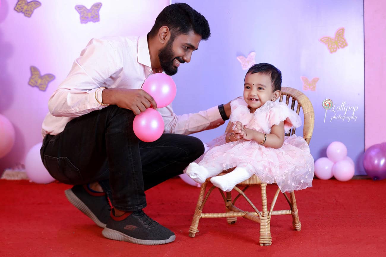 Joyful father playing with his little daughter, who smiles brightly while seated on a rattan chair amidst a whimsical birthday setup in Chennai, captured by Lollipop Photography.